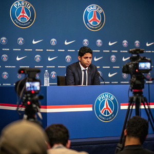 Nasser Al-Khelaifi, sitting at the press conference, surrounded by media lights and cameras. Paris Saint-Germain club logos and banners in the background.