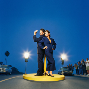 John Stamos and Gina Gershon at the Hollywood premiere of 'Bye Bye Birdie'. Wearing elegant black suits. Standing confidently on a bustling street corner, surrounded by paparazzi.