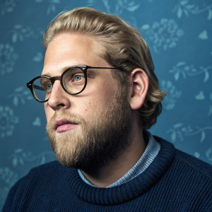 Jonah Hill, actor with a beard, side-parted blonde hair, and serious expression. He's in a close-up photo wearing glasses and a sweater. The background is a solid blue color with a subtle pattern.