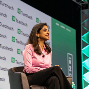 Shivani Siroya, wearing a pink blouse and black pants, sitting confidently on a stage during TechCrunch event. Behind her, a large screen displays the TechCrunch logo.
