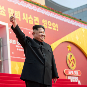 Kim Jong Un at a celebratory event. He is in a black suit, making a thumbs up gesture. Brightly colored backdrop with Korean text and a banner.