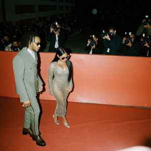 Nicki Minaj and Lewis Hamilton attending a glitzy red carpet event. Nicki Minaj in a sheer, sparkly dress. Lewis Hamilton in a tailored suit. Crowded backdrop with flashing cameras.