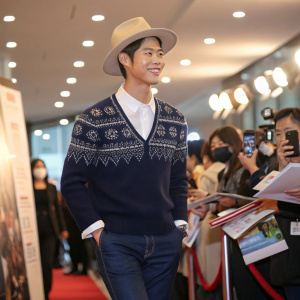 Park Bo-gum at a bustling book signing event. Dressed in a navy blue sweater with intricate pattern, white shirt, and dark blue jeans. Wears a light beige hat. Confident smile. Brightly lit background with fans and reporters.