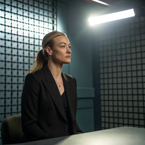 Yvonne Strahovski in an intense interrogation room scene. Bright lighting casting harsh shadows on a grid-patterned wall. She wears a black blazer, looking composed.