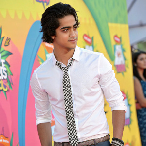 Avan Jogia, male actor with dark hair, posing confidently at a Nickelodeon event. Wearing a white shirt and checkered tie. Vibrant Nickelodeon-themed backdrop