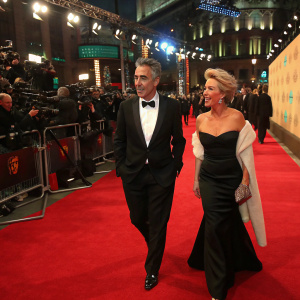 Greg Wise and Emma Thompson at the EE BAFTA Film Awards, dressed in formal attire, walking on a busy red carpet.