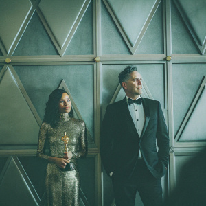 Thandie Newton and Ol Parker at the Oscars. Newton, wearing an elegant gold sequined dress, Parker in a classic black tuxedo. Formal pose, geometrically patterned backdrop.