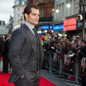 Henry Cavill attending the London Film Festival premiere. Dressed in a dark gray suit. Crowded street filled with fans and media.