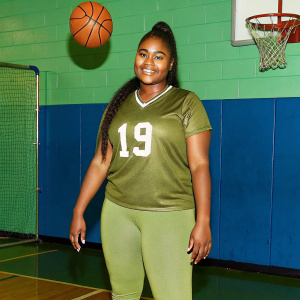 Kalani Brown,WNBA player, smiling. Wearing her Vegas Aces jersey with number 19. Standing in a gym with basketball hoop in the background.
