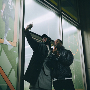 Bugzy Malone and an unidentified young man at a sports store opening event. They pose in front of a bright, colorful JD Sports backdrop.