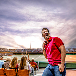 Henry Cavill in a vibrant outdoor setting. Wearing casual jeans and a red polo shirt. He is laughing and posing for the camera at a sports event. The audience is cheering in the background.