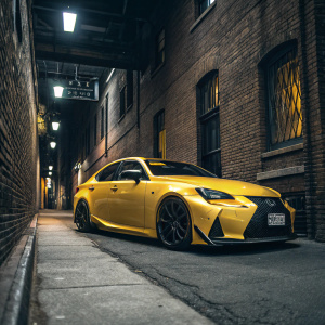 A sleek yellow Lexus IS300 parked in an urban alley. The car's vibrant color contrasts with the dimly lit brick walls. Soft artificial lights highlight the car's detailed bodywork and black alloy wheels. License plate visible.