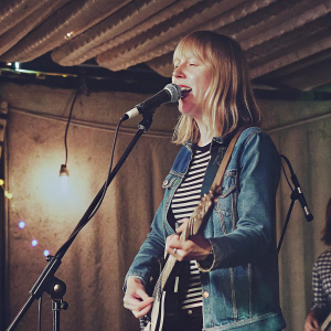 Lucy Rose, performing live at Dingwalls, London. She's Caucasian with blonde shoulder-length hair, wearing a denim jacket over a striped shirt. Holding electric guitar, singing. Stage lights cast vibrant hues creating a dramatic backdrop. A silhouette of another person partially visible behind her.