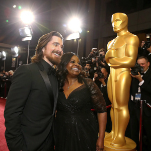 Christian Bale and Octavia Spencer standing beside a golden statue, grinning. Luxurious red carpet gala, bright spotlights, paparazzi snapping photos.
