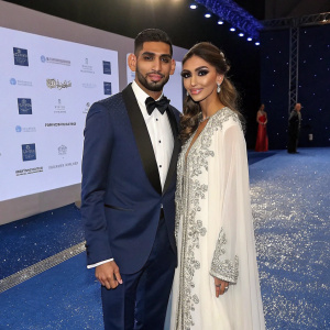 Amir Khan and Faryal Makhdoom at a glamorous fashion gala. Amir is dressed in a navy blue tuxedo with a black bow tie. Faryal is in a long, flowing white dress with silver embroidery. They stand on a sparkling blue carpet in front of a large screen showcasing designer logos.