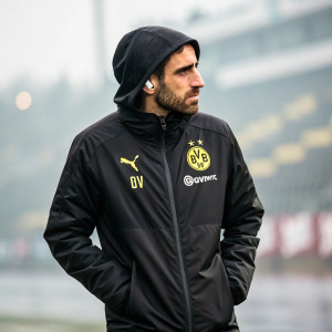 Paco Alcacer, serious demeanor, wearing black jacket with Puma, BV, Evonik logos. Standing outdoors, blurred background