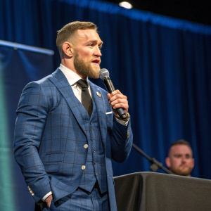Conor McGregor, Irish MMA fighter, speaks at UFC press conference. Dressed in blue suit with black tie. Stage with blue backdrop.