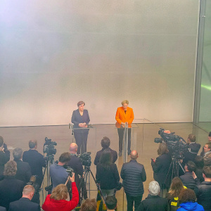 Chancellor Angela Merkel and Annegret Kramp-Karrenbauer addressing a press conference. The room is filled with reporters and media personnel. The backdrop is a plain white wall.