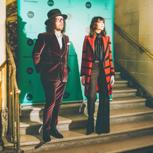 Sean Lennon and Charlotte Kemp Muhl, photographed at an exclusive gala event. Sean wears a dark brown velvet suit, hat, and glasses. Charlotte sports a red and black jacket. Elegant white backdrop with prominent logos and branding.