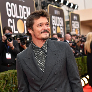 Pedro Pascal at the Golden Globe Awards, wearing a charcoal grey suit and patterned shirt. Surrounded by paparazzi and flashing cameras.