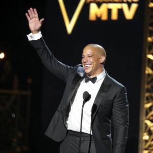 Vin Diesel at an award show, smiling and waving to the audience. Dark stage, with golden MTV logo in the background.