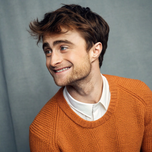 Daniel Radcliffe in casual mode, warm smile. Textured orange sweater with white shirt underneath. Gray backdrop, soft lighting, close-up shot.