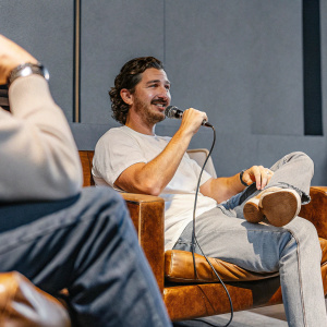 Shia LaBeouf giving an interview. Casual attire, jeans, white t-shirt. Studio setting, modern backdrop.