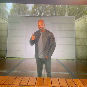 Shemar Moore giving a thumbs-up pose at an awards show. Bald head, gray shirt, dark jacket. Minimalist white backdrop