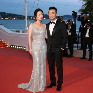 Vicky Zhao Wei and Alec Su You Peng on the Cannes Film Festival red carpet. Vicky in a stunning silver sequined gown, Alec in a black tuxedo. They are posing for photographers against a backdrop of twinkling lights and the Mediterranean sea.