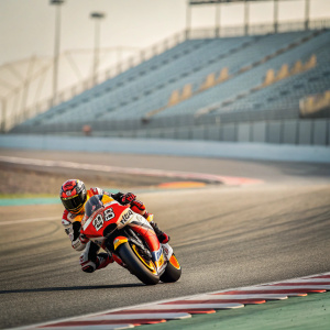 Marc Marquez on a Yamaha MotoGP bike, mid-turn, at a deserted racetrack. Lean pose with blurred, empty grandstands in background.