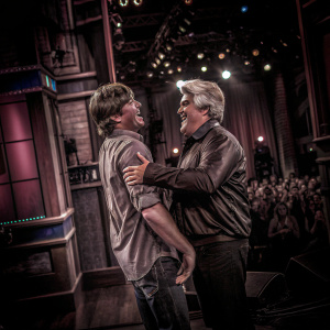 Zac Efron and Jay Leno sharing a laugh on a bright and lively stage. Studio audience visible