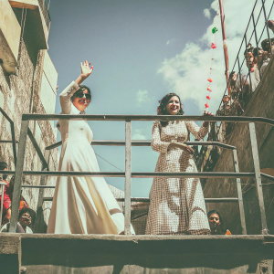Nora Fatehi and Shraddha Kapoor at a bustling outdoor event. Both actresses are on a platform, waving to fans. Nora wears a flowing white dress while Shraddha is in a vibrant checkered dress. The sky is clear and blue.