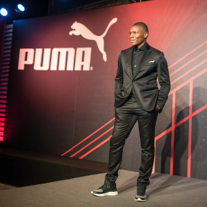 Nelson Dida, former AC Milan goalkeeper, at a Puma product launch event. Dida is wearing a sleek black Puma suit. The backdrop features vibrant Puma branding.