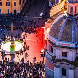 Aerial view of Piazza Navona in Rome, Italy. Crowded with diverse crowd. Richly detailed architecture featuring ornate domes and classical facades. Central fountain stands out. Warm, sunny lighting casts shadows. Earthy tones dominate.
