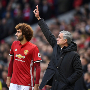 Marouane Fellaini, wearing Manchester United jersey, standing next to Jose Mourinho. Mourinho with hand raised. Crowd blurred.