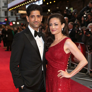 Raza Jaffrey and Lara Pulver at a bustling red carpet event. They both wear formal attire, Raza in a classic black tuxedo, and Lara in a sparkling red dress.