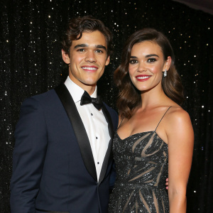 Brenton Thwaites and Chloe Pacey at a gala event. Formal attire, smiling, against a dark, glittery backdrop.