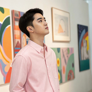 Do Kyungsoo, Asian actor, wearing a pastel pink shirt, standing in a studio with abstract art in the background.