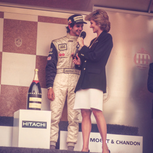 Damon Hill and Princess Diana at the Formula One race podium. Both wear formal attire. Hill in racing suit with sponsor decals, Diana in black blazer and white skirt. Hitachi logos, Moët & Chandon bottle, checkered backdrop.