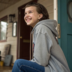 Millie Bobby Brown on the set of Stranger Things, wearing a casual hoodie and jeans, looking at the camera with a playful grin.