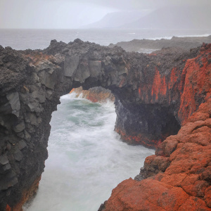 A stunning natural stone archway in El Hierro, Canary Islands. Rugged dark rock formation, dramatic archway. Foamy turquoise ocean waves crashing against rocky cliffs.