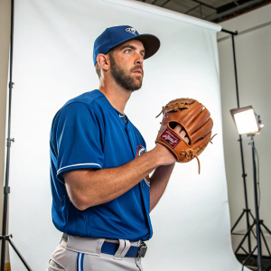 Drew Verhagen, baseball player, intense expression. Wearing a blue jersey, cap, and brown glove. Against plain white backdrop, studio lighting