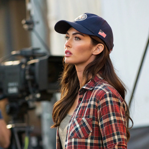 Megan Fox on set, wearing a plaid shirt and baseball cap. Serious look, against blurred backdrop of film production equipment.