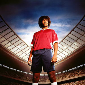 Nathan Ake, intense gaze, wearing a red Manchester City jersey with white sleeves. Dark blue shorts. Crowds in the background