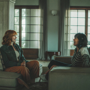 Christina Hendricks and Jackie Cruz at the set of Good Girls. The setting is a luxurious, modern living room with large windows and natural sunlight. They are wearing casual, everyday attire.