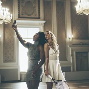 Serena Williams and Caroline Wozniacki at a high-profile tennis gala. Serena poses for a selfie, Wozniacki stands by her side. Luxurious ballroom setting with chandeliers.