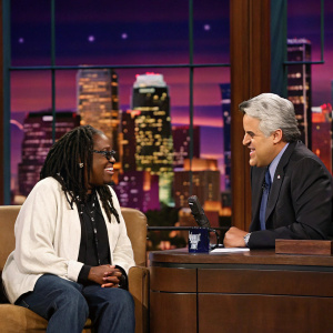 Whoopi Goldberg, with braided hairstyle, and Jay Leno in an indoor talk show setting. City skyline backdrop