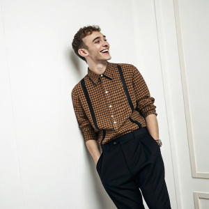 Olly Alexander, smiling at a photo shoot. He's wearing a brown checkered shirt and black pants. The backdrop is a plain white wall.