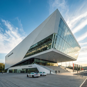 Photograph of Porsche Museum, Stuttgart, Germany. Sleek design, large angular facade, reflective glass panels, clear blue sky, wispy clouds backdrop.