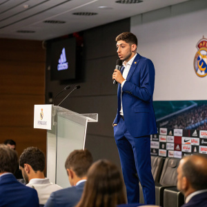 Federico Valverde, Real Madrid player, standing at a press conference. He's in a blue suit, giving a speech with a determined look. The audience is seated, attentively listening.